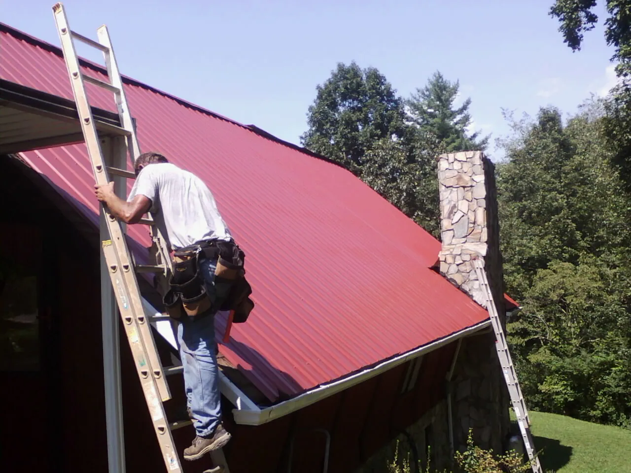 Roofer on a ladder beside a red metal roof and stone chimney — Soffit & Fascia Repair by Roofing Brothers in Kittredge